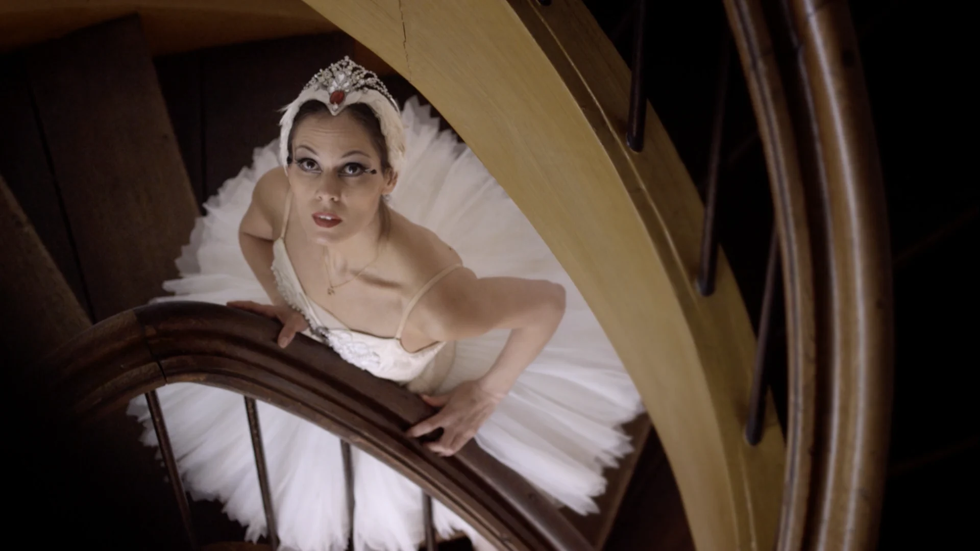 A ballerina in a tiara looks up from a curved wooden banister