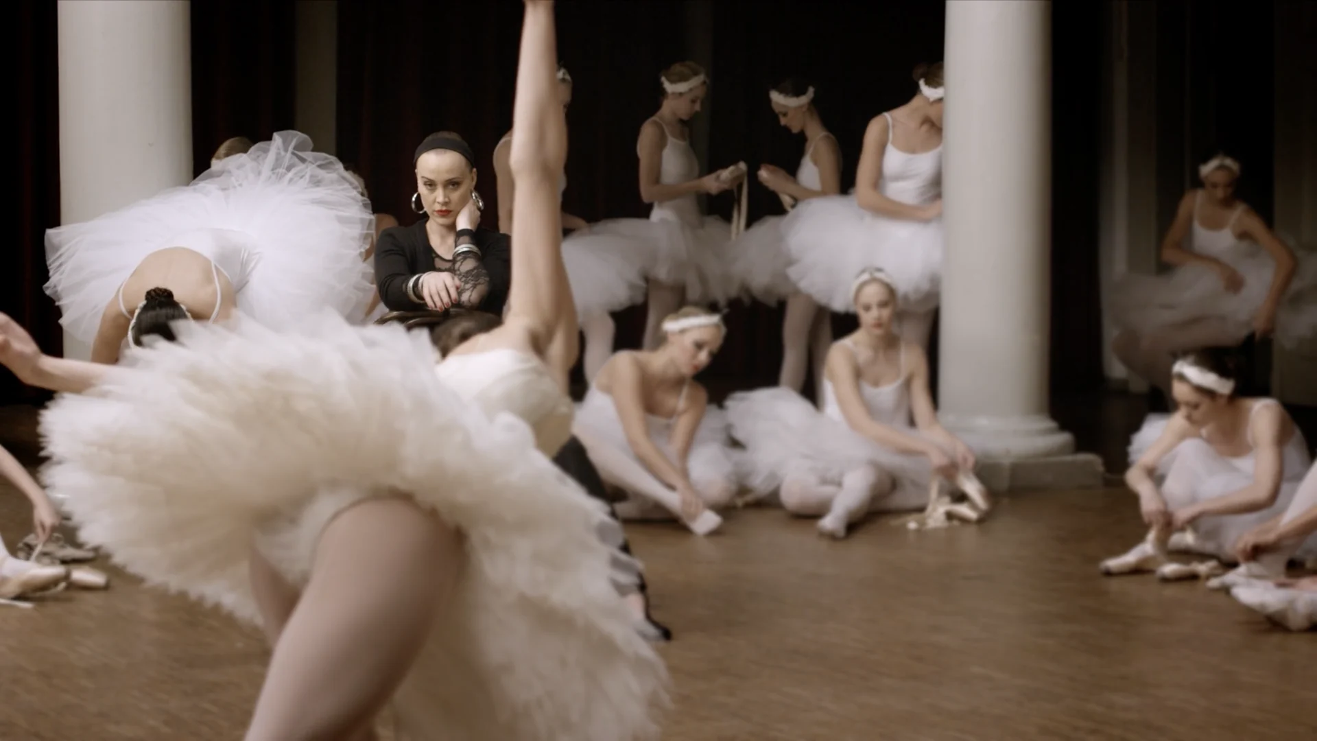A dancer in a tutu bends back in the foreground, the corps de ballet tying their shoes behind her