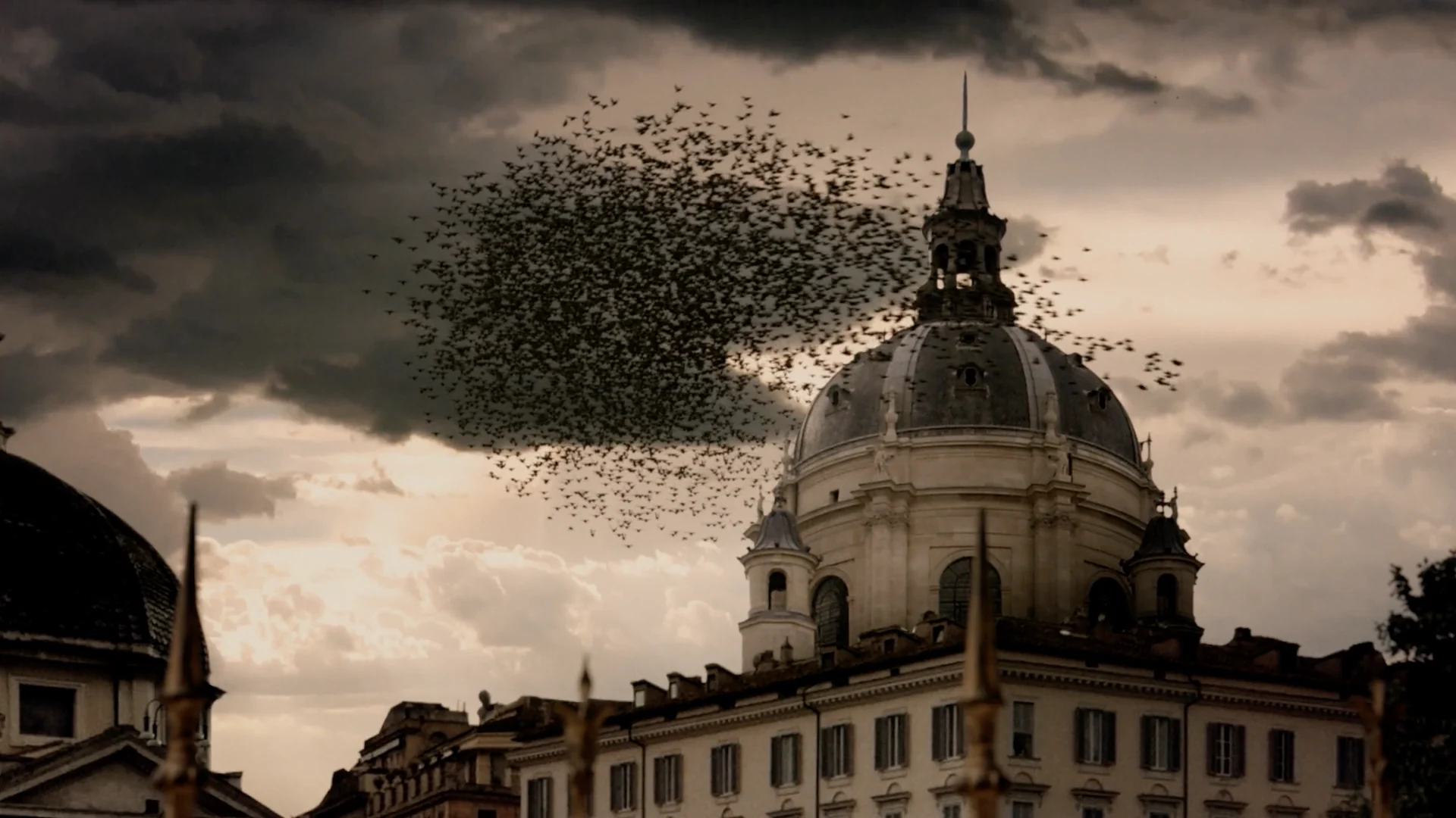 A huge flock of birds swirls past a domed church under a dark sky