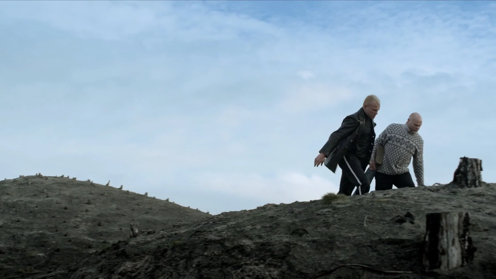 Two men walk over the crest of a dune under a pale sky, in The Good Life