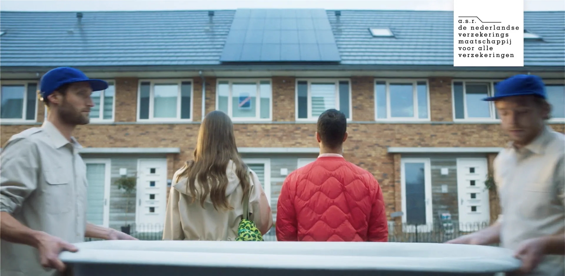 Two removal men carry a bathtub past a couple outside a brick house with solar panels on the roof