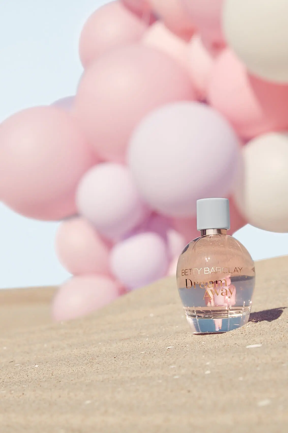A bottle of Dream Away stands in rippled sand in front of pink balloons