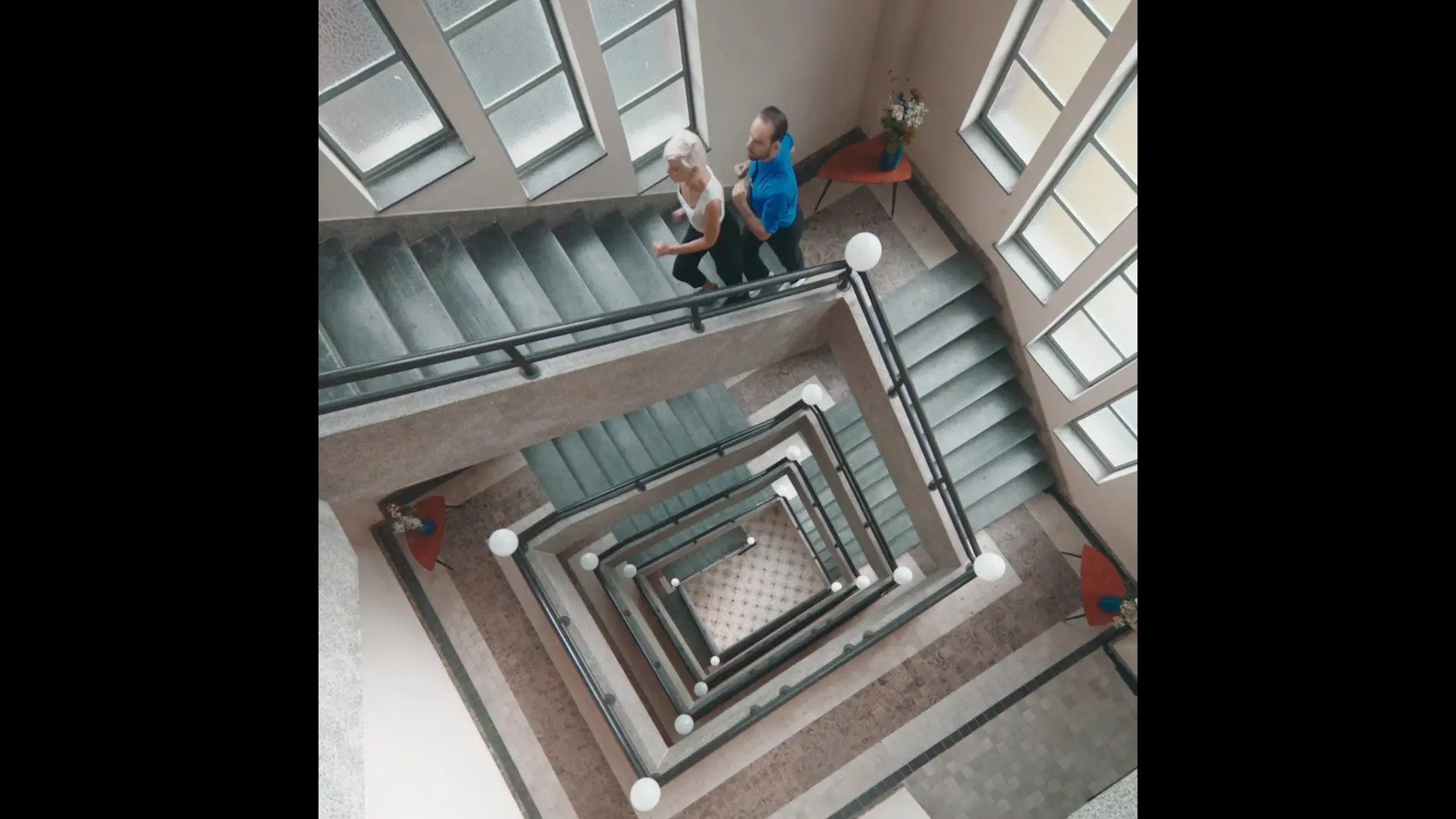 Still from the De Staat music video Round, two people running up a square stairwell seen from above