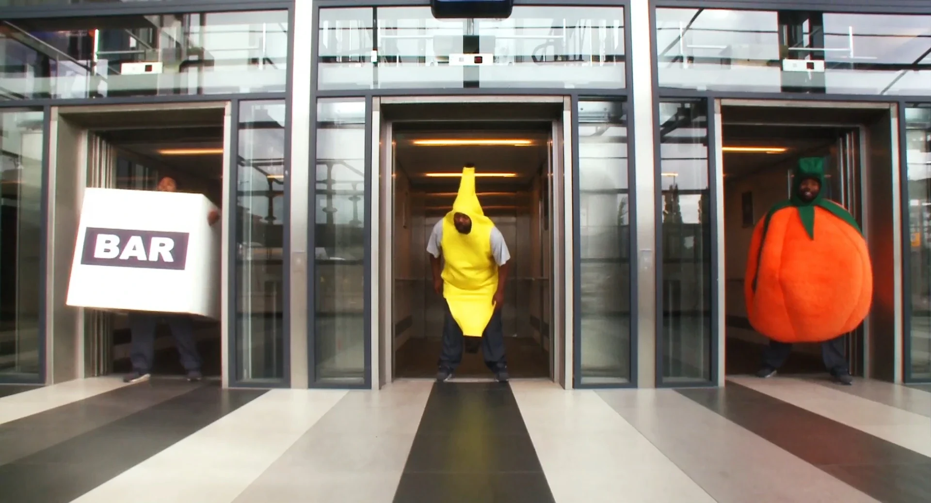 A man in a banana suit, one in an orange suit and one dressed as a box marked BAR step out of three lifts