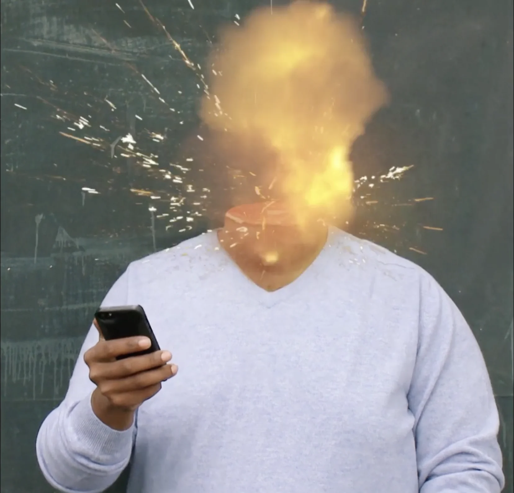 A man in a grey jumper holds his phone while his head explodes in flames