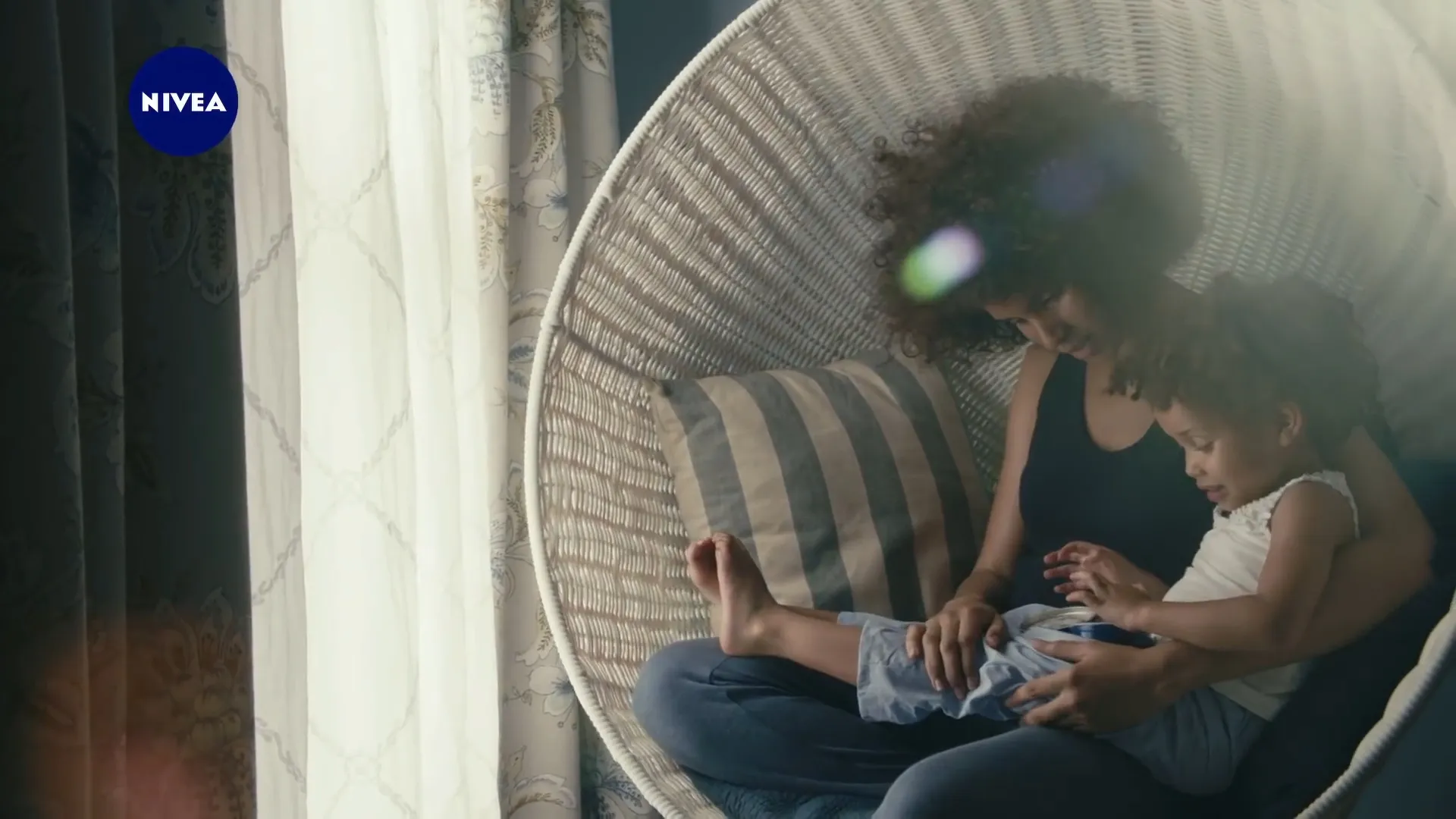 A mother and her daughter in a hanging wicker chair by a window, from the Nivea Crème commercial