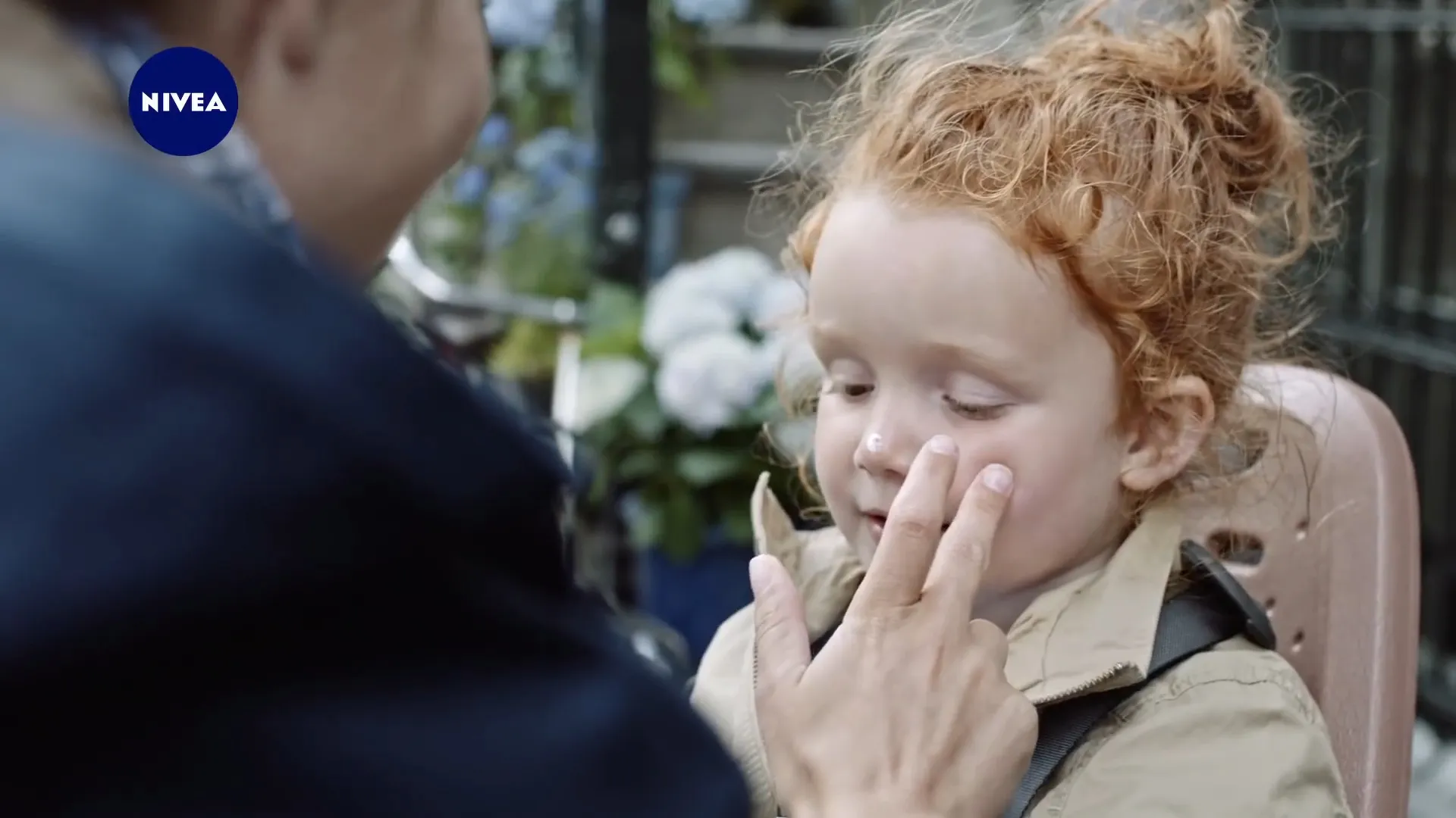 A red haired girl on a bike seat gets cream on her cheek, canal houses and flowers behind her
