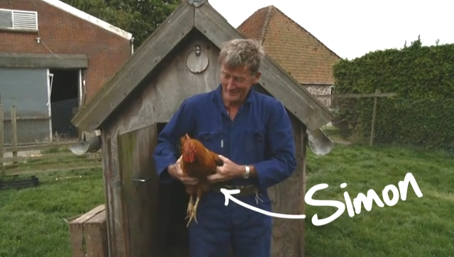 A farmer in blue overalls holds his rooster Simon outside the coop