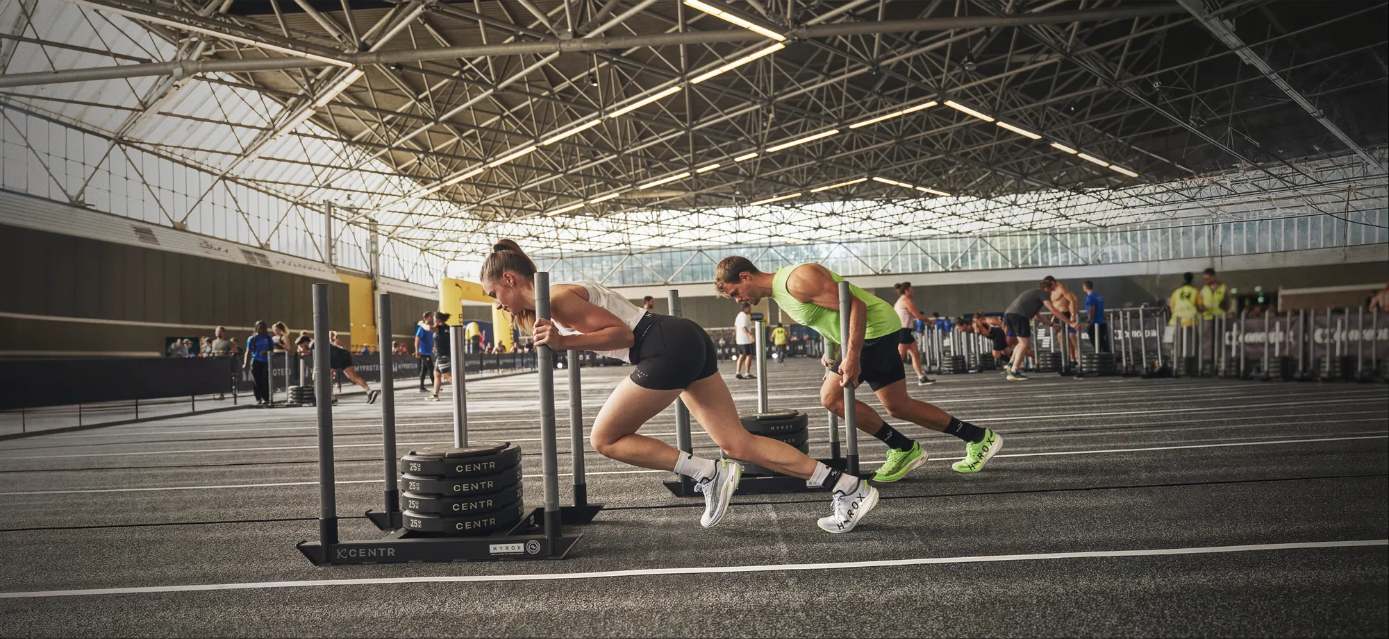 Two athletes pushing sleds under the roof of the RAI hall