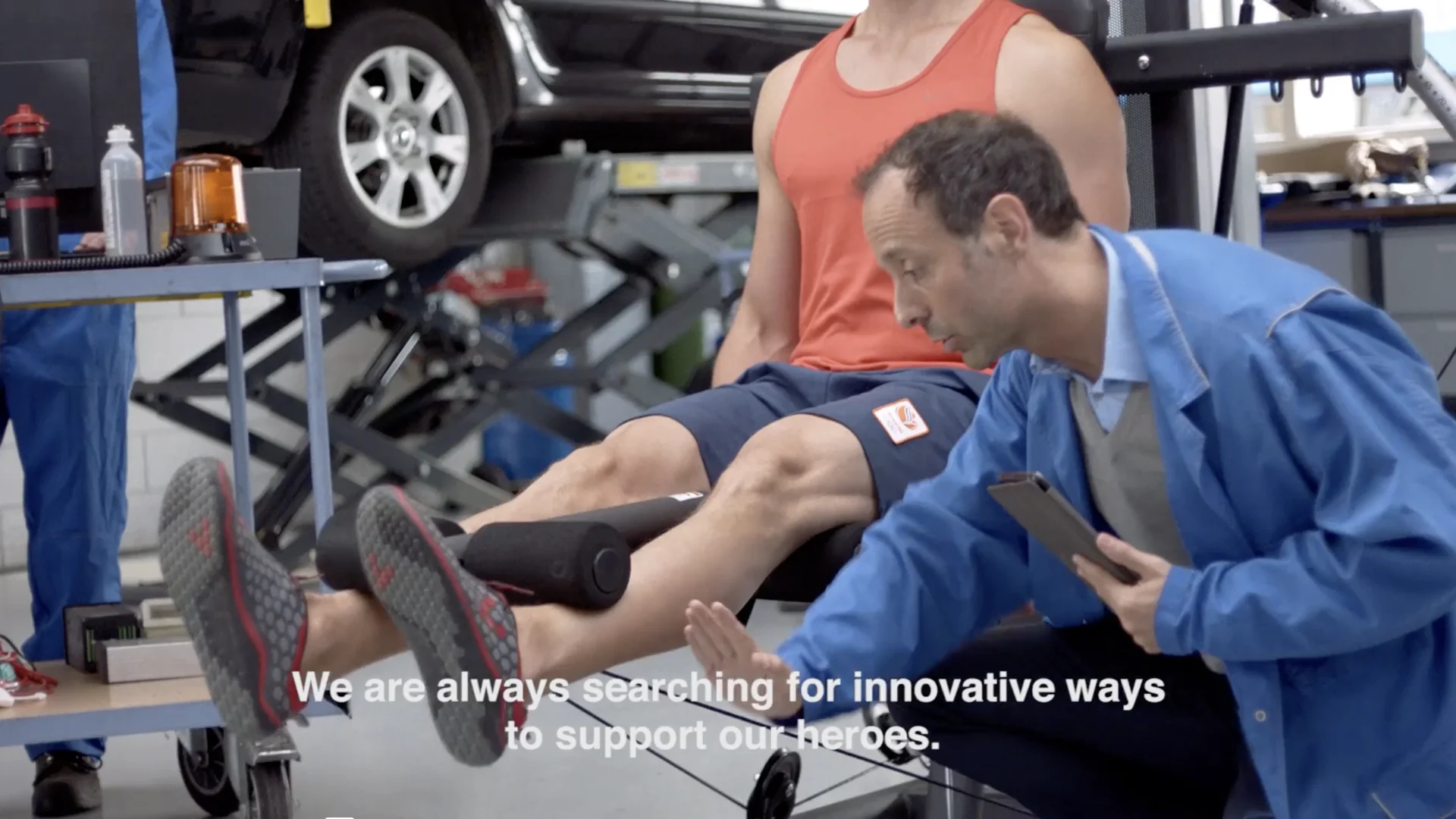 An engineer checks an athlete on a leg press set up between the cars in the workshop