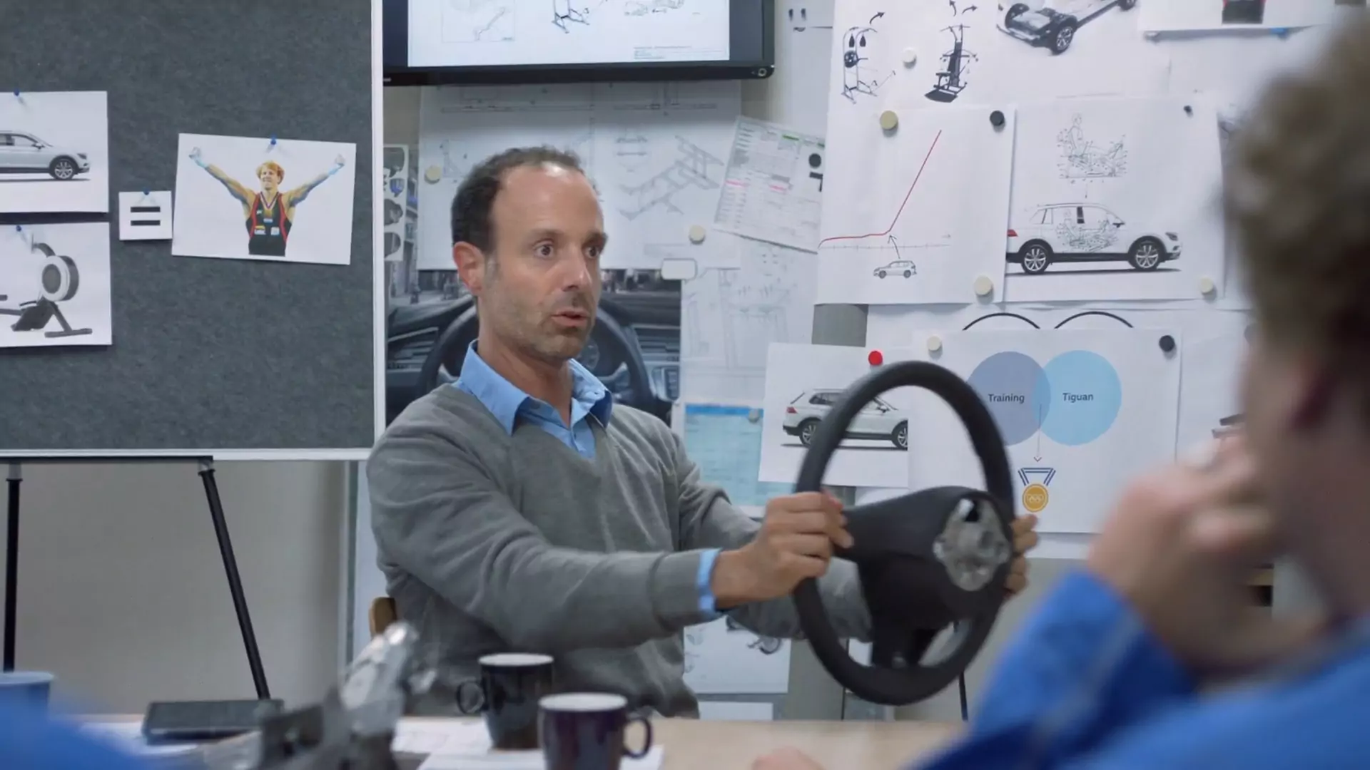 An engineer holds up a steering wheel in front of a wall of sketches