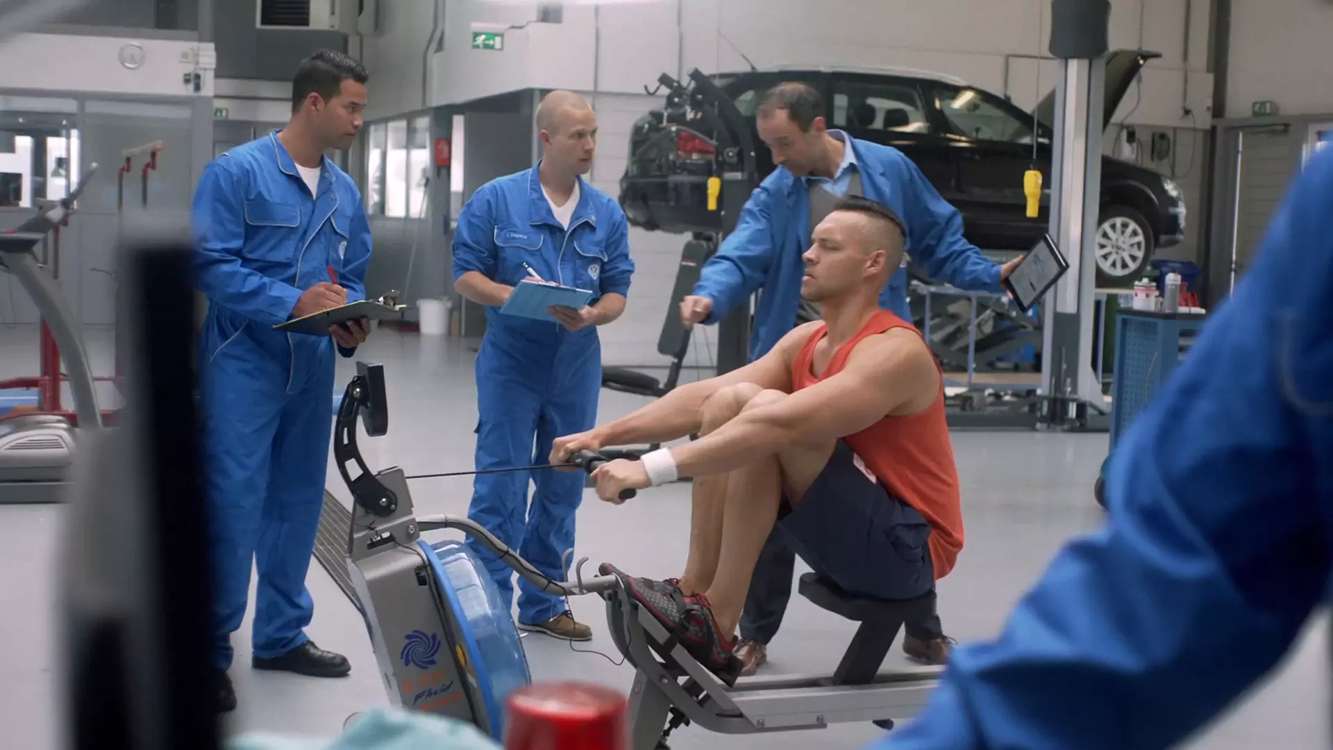 Mechanics in blue overalls take notes while an athlete trains on a rowing machine in the workshop
